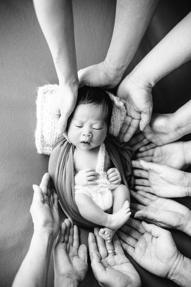 Sanfte Neugeborenenfotografie, Baby mit Familienhänden, schwarz-weiß, liebevolles Babyfoto, Baby von mehreren Händen umgeben