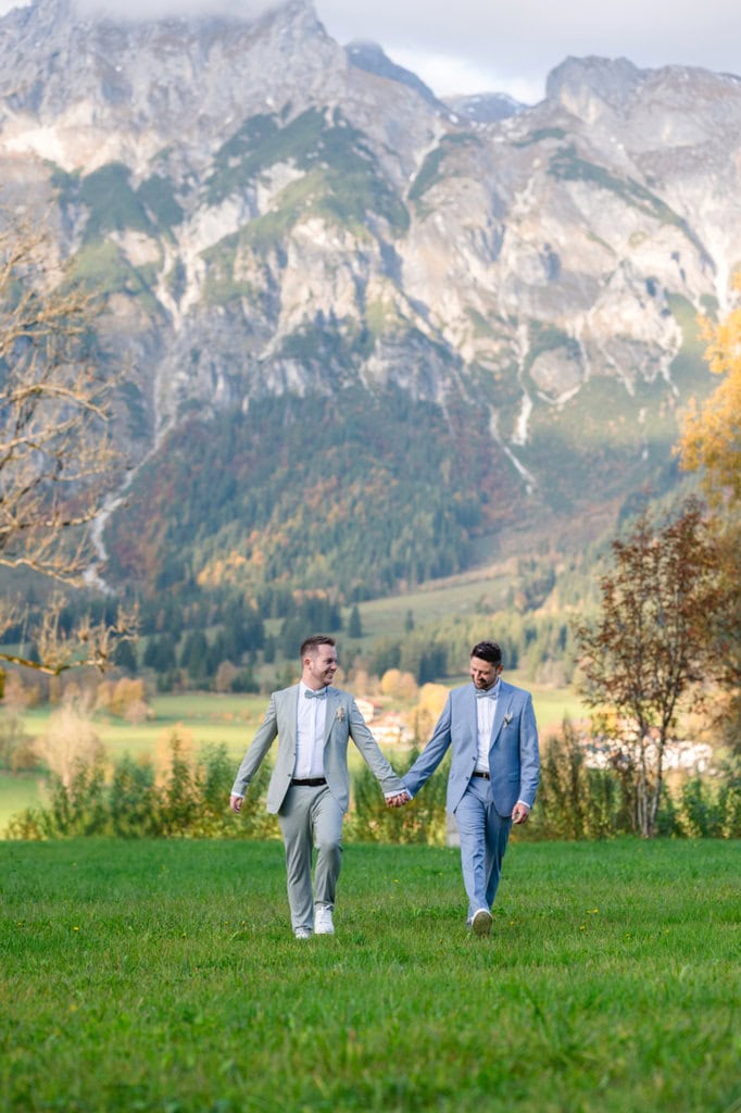 Männer-Hochzeitspaar spaziert Hand in Hand über eine grüne Wiese.