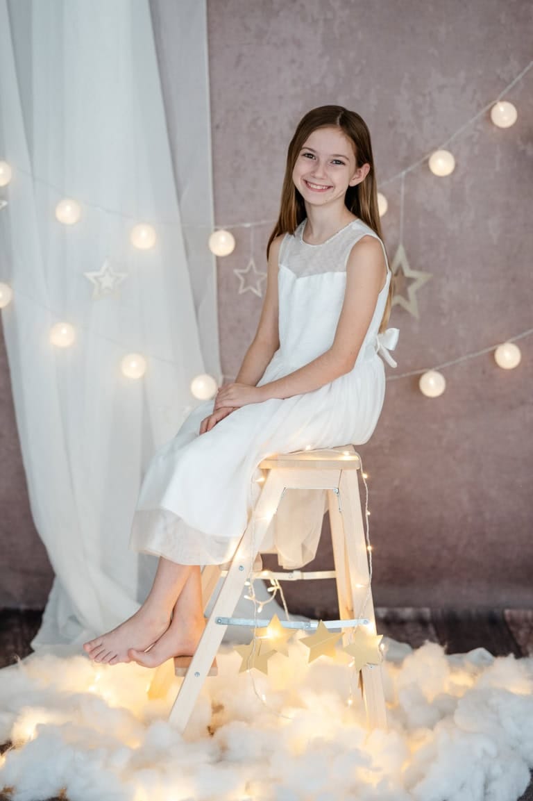 Lächelndes Mädchen in weißem Kleid sitzt auf einem Holzstuhl vor einer festlichen Dekoration mit Lichterketten und Sternen.