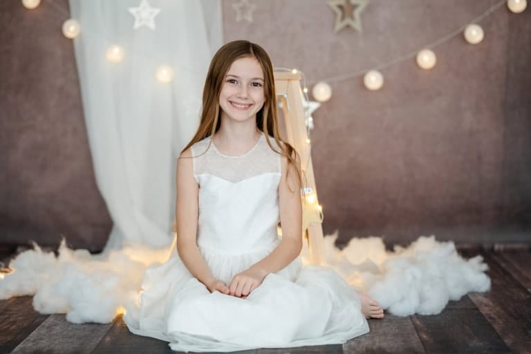 Lächelndes Mädchen in weißem Kleid sitzt auf einem weißen Teppich vor weihnachtlicher Dekoration.