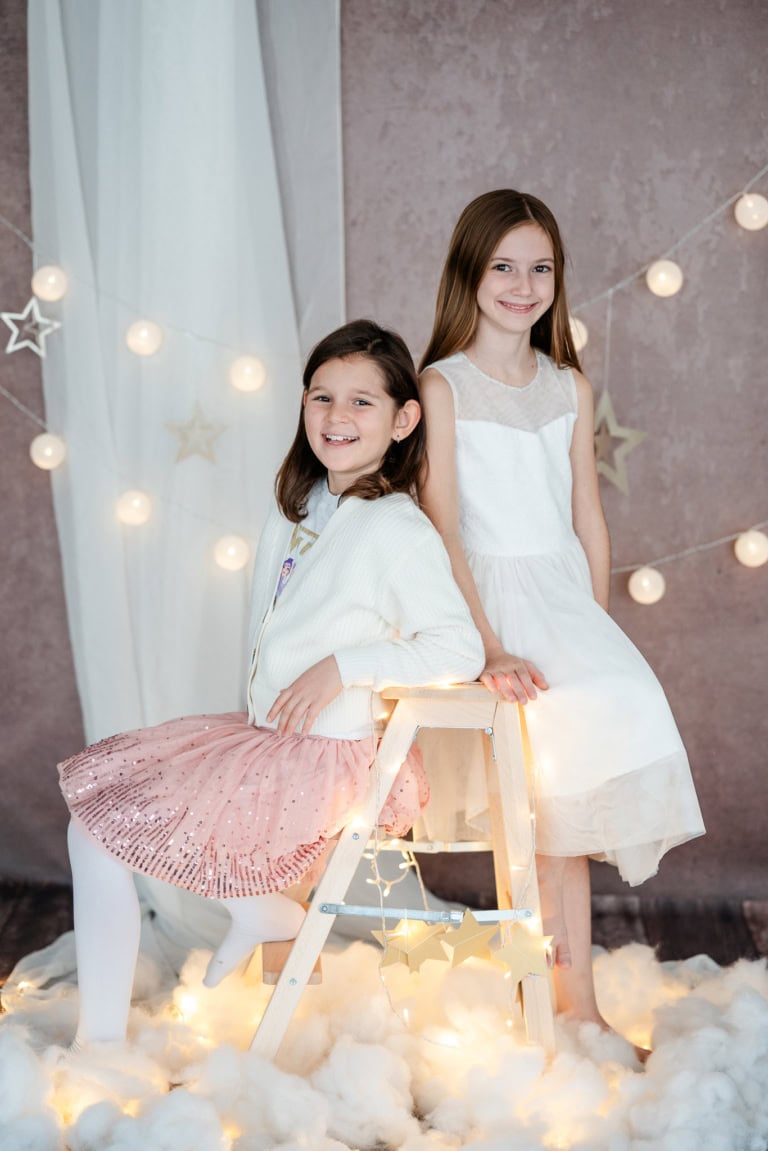 Zwei lachende Mädchen in weißen Kleidern bei einem festlichen Fotoshooting mit Lichterketten und Sternen-Dekoration.