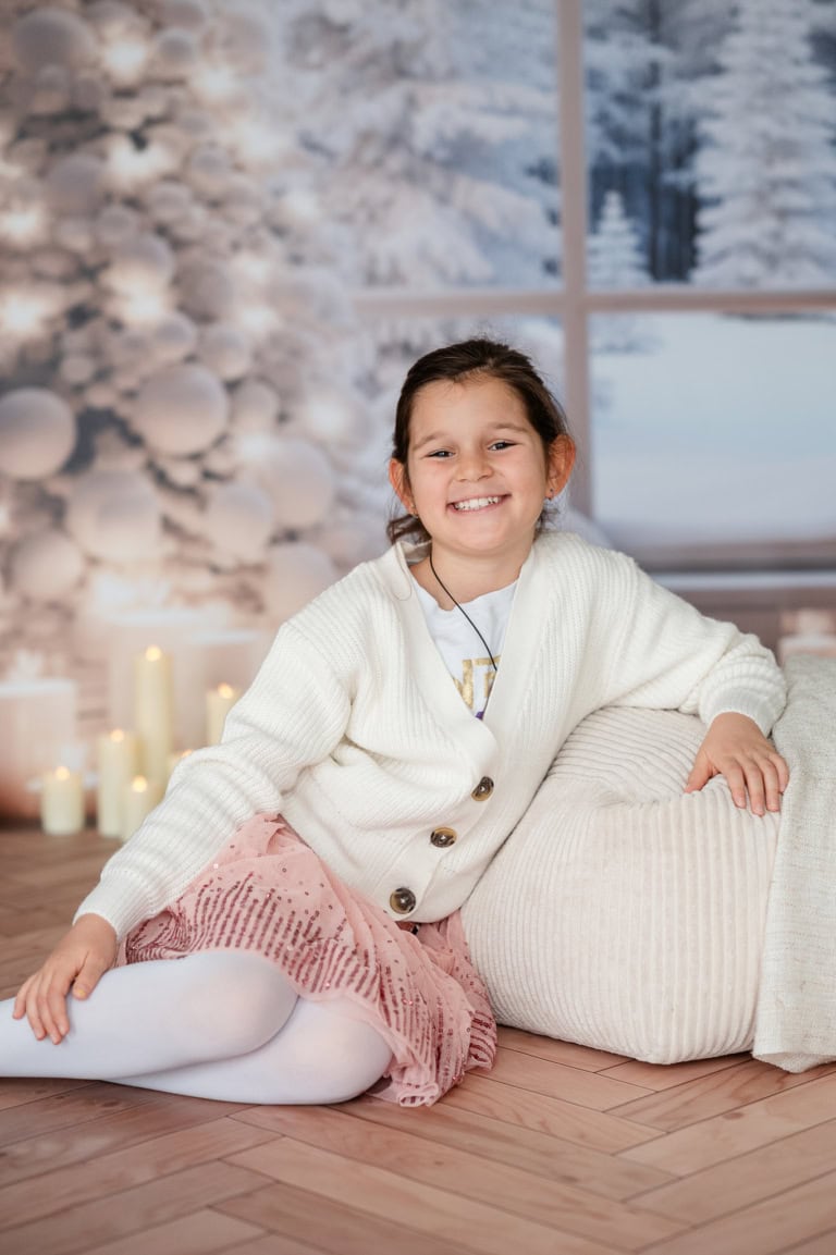 Fröhliches Mädchen sitzt auf einem Teppich vor einem Weihnachtsbaum mit Kerzen, Winterlandschaft im Fenster im Hintergrund.