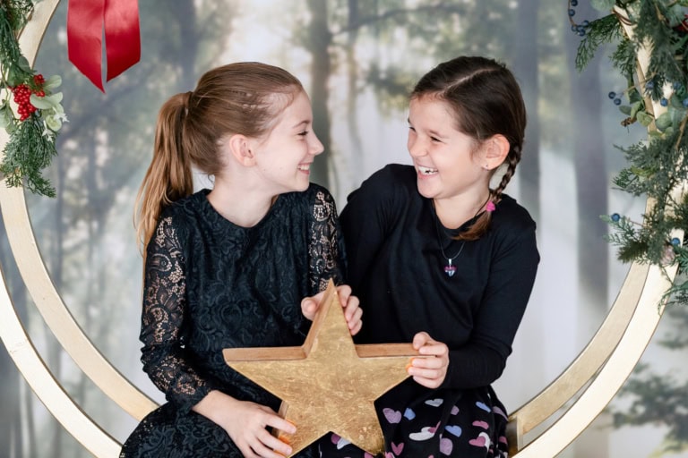Zwei lachende Mädchen sitzen auf einem Weihnachtsring, halten einen goldenen Stern und schauen sich fröhlich an.