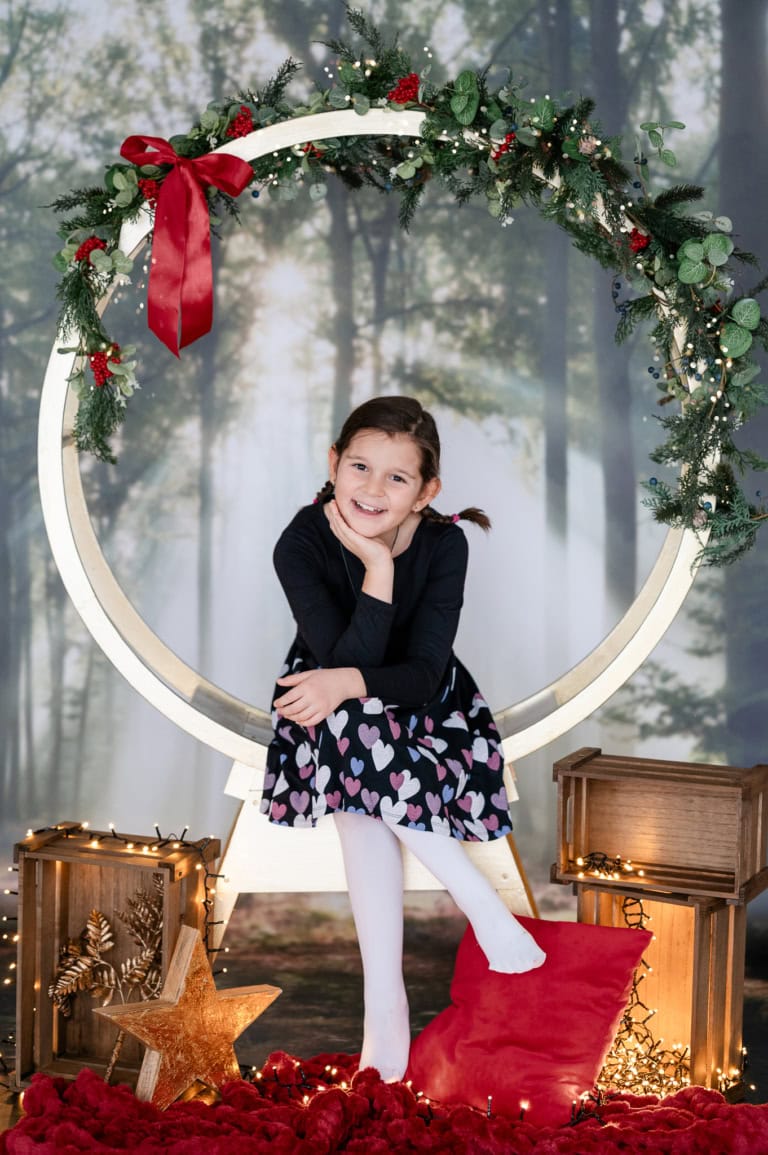 Junge Mädchen sitzt vor einem festlich geschmückten Rahmen mit Weihnachtsdeko, lächelt und posiert für ein Weihnachtsfoto.