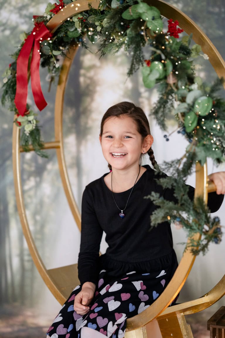Fröhliches Mädchen sitzt in einem goldenen Rahmen mit Weihnachtsdekoration, lächelt und trägt ein schwarzes Oberteil und eine Hose mit Herzmustern.