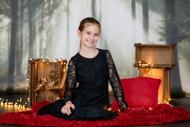 Junge Frau in schwarzem Spitzenkleid sitzt auf rotem Teppich vor weihnachtlicher Kulisse mit Lichterketten und Dekoration.